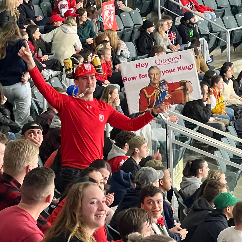 A lively crowd at the Carr-Harris Challenge Cup hockey game. In the foreground, a fan dressed in red, wearing a beer helmet, stands with arms raised, holding a sign that reads, “Hey Queen’s! Bow for your King!” featuring an image of King Charles III. Other fans in the stands, many wearing red and black, cheer and engage in the spirited rivalry between teams. The atmosphere is energetic, with spectators reacting enthusiastically to the game.