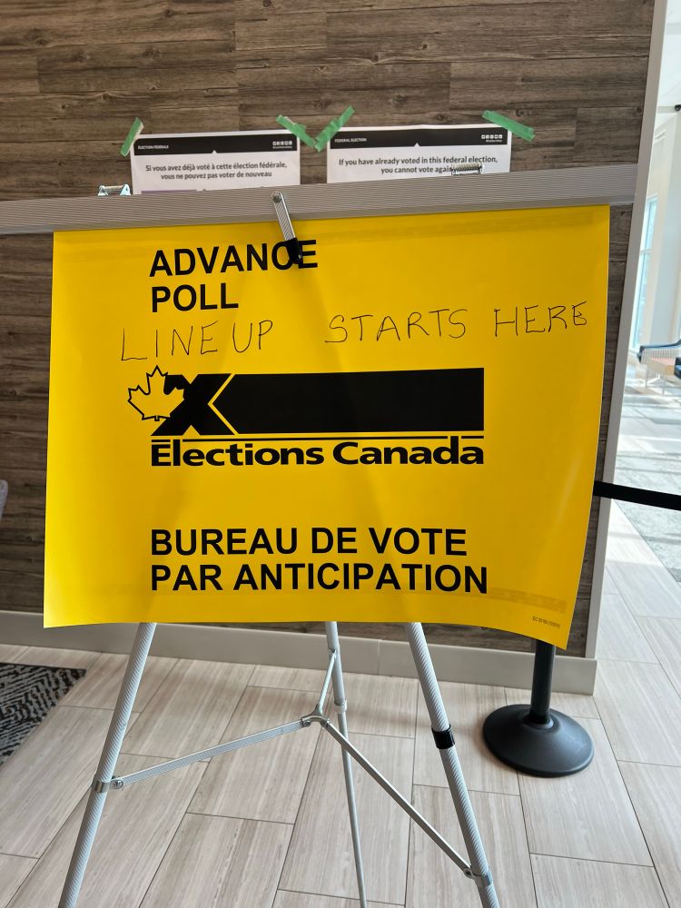 A yellow Elections Canada sign indicating the location of an advance polling station. The sign reads “Advance Poll – Lineup Starts Here” in both printed and handwritten text, and “Bureau de vote par anticipation” in French. The sign is displayed on a tripod stand in a public indoor space.
