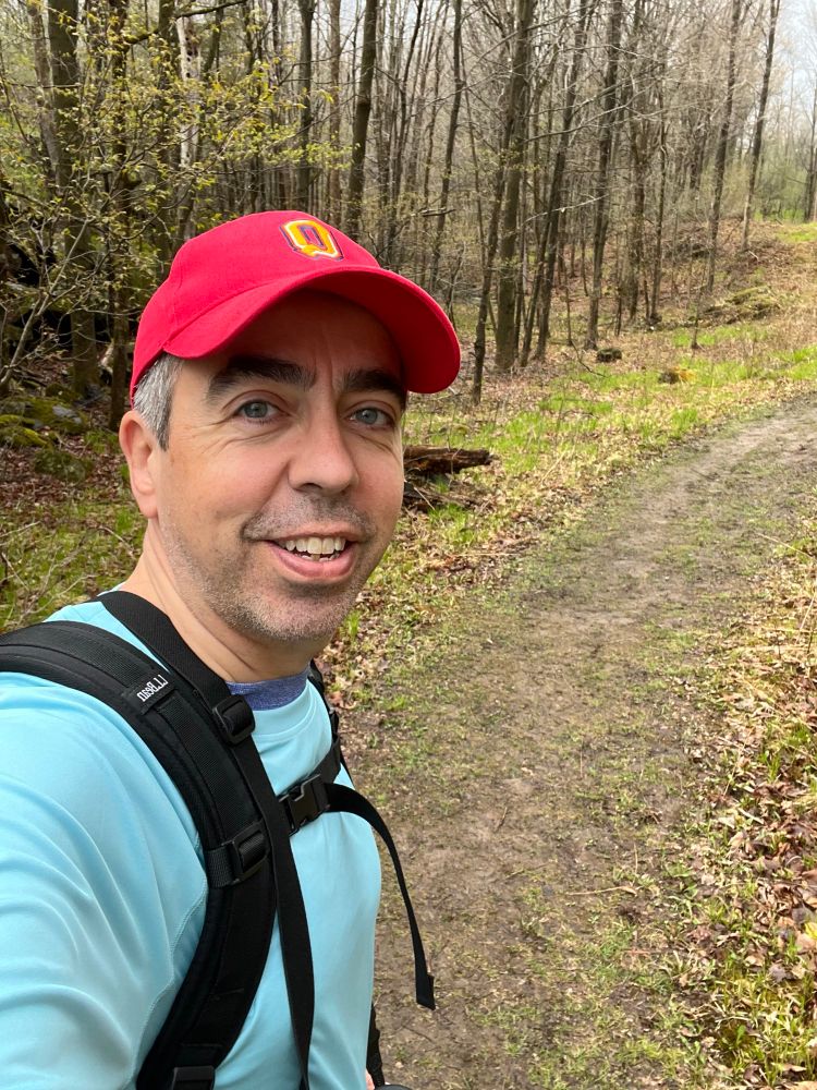A white man wearing a red Queen’s University cap, light blue shirt, and black backpack takes a selfie on a forest trail in Frontenac Provincial Park. The background features early spring trees with fresh green growth and a winding dirt path.