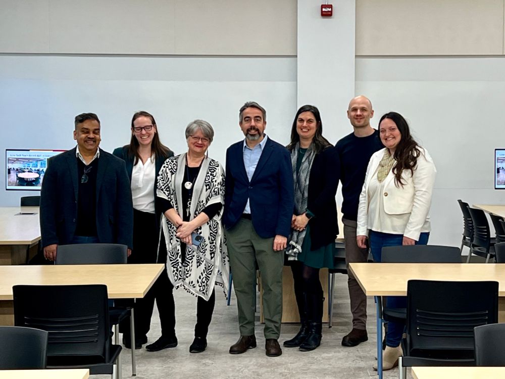 A group of seven people standing together in Dunning 10, a newly opened learning space. They are dressed in business casual attire and smiling at the camera. The background features modern classroom furniture, including tables, chairs, and screens displaying images of the space. The setting reflects a collaborative and welcoming environment for teaching and learning.