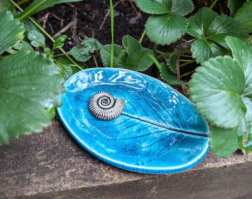 Turquoise blue pin tray with a real cherry tree leaf pressed into it for the pattern in horizontal position upon an old railway sleeper with strawberry leaves surrounding it. There's a hand crafted ammonite at one end. 