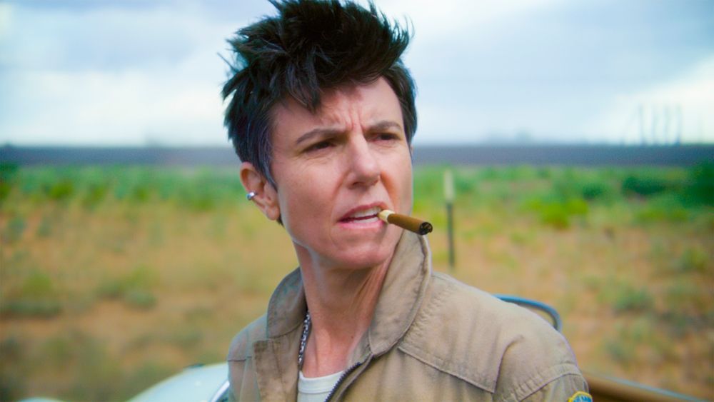 Tig Notaro in an engineer's jumpsuit, squinting in the sunlight with a cigarillo clamped in her teeth. Her hair is tousled and she is wearing a silver ring through her conch.