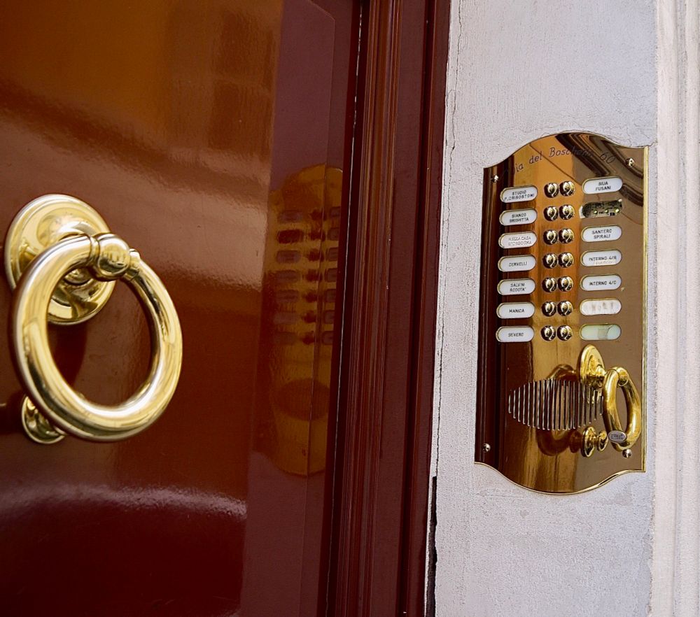 Goldener Ring als Türgriff an einer braunen glänzenden Tür, daneben an der Wand ein goldenes Klingelschild. 