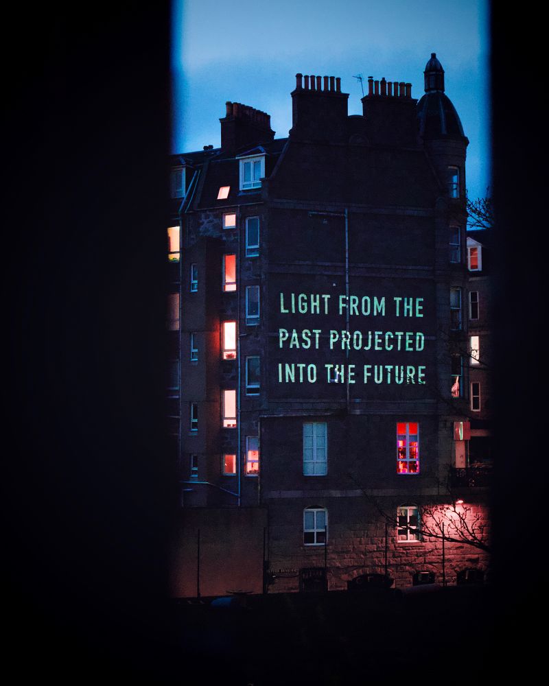 A tenement building with “light from the past projected into the future” projected on the side. 