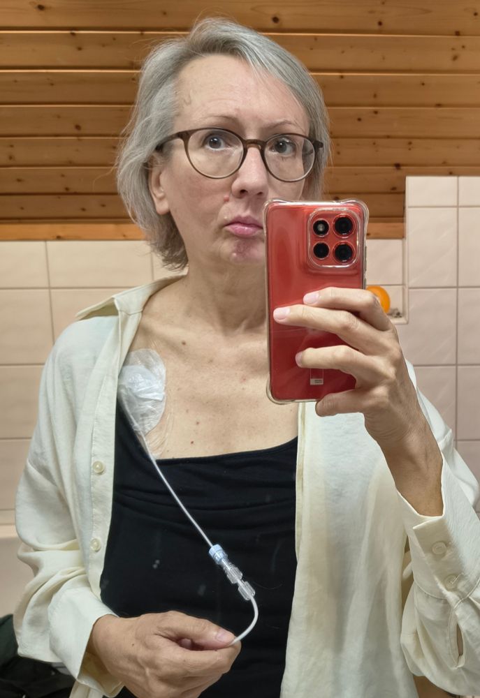 Die Account-Inhaberin schaut aus einem Badezimmerspiegel heraus ins Leere. Sie hält ein Handy in der Hand, das einen kleinen Teil ihres Gesichts verdeckt. Ihr Haar ist grau und relativ kurz, und sie ist Brillenträgerin. Sie trägt ein schwarzes Top und eine helle Bluse. Unter ihrem Schlüsselbein ist ein großes Pflaster, das einen Port verdeckt. Ein dünner Schlauch für parenterale Ernährung ist an den Port angeschlossen.