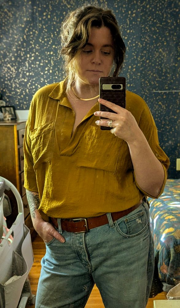 A gender fluid person in jeans, a brown belt, mustard yellow loose top, and gold necklace taking a selfie in a mirror. They have long dark brown hair with a blonde streak pulled back in a ponytail.