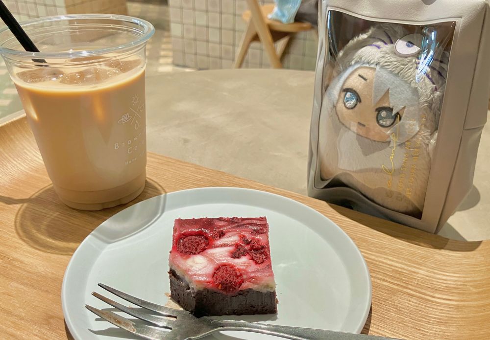 テーブルの上のトレイ、左からカフェラテ、ひとくち分欠けたラズベリーとクリームチーズのブラウニー、窓付きケースに入ったモンがく。