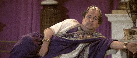 Dom DeLuise as a Roman emperor wearing a purple toga with gold trim over a white tunic and a gold laurel lounging on a bench saying “Nice, not thrilling, but nice”