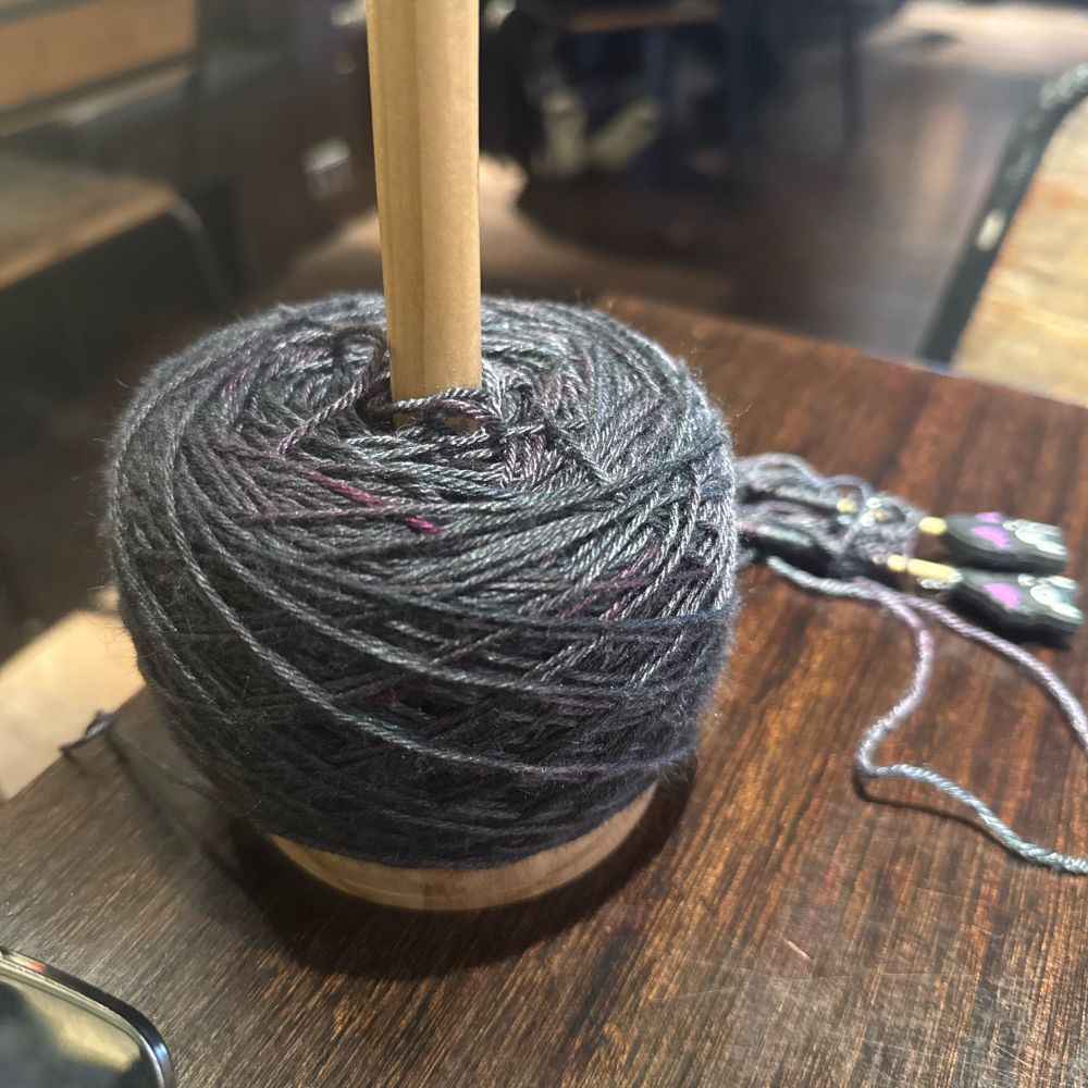 Photo of a cake of yarn on a spinner set on a table. It’s a beautifully soft silk merino blend and looks almost oil bit like an oil slick - silvery grey dominates but with lovely shades of purple and blue in the spin. 