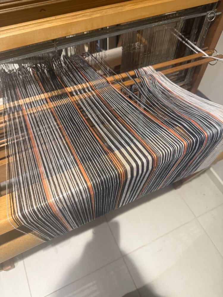 A photo from the back of my loom showing my warp  of black and white cotton threads in various patterns with light orange pattern breaks. It’s would onto the back beam and about 4/5 threaded through the heddles. 