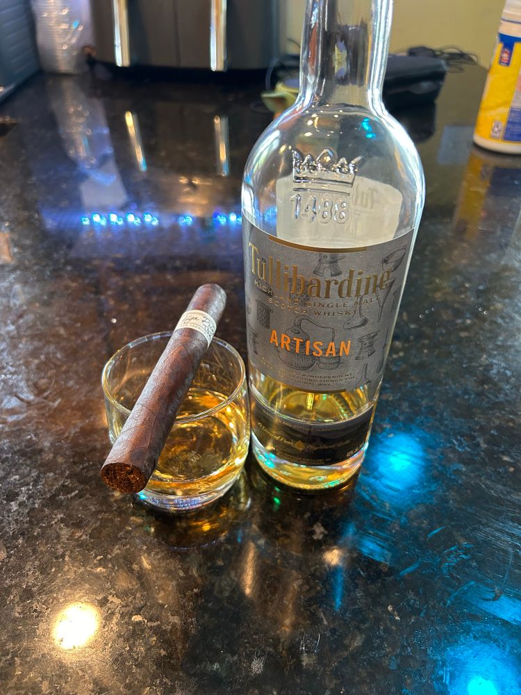 On a black granite counter top sits a bottle of inexpensive single-malt scotch next to a glass with a generous pour. Atop the glass balances a Liga Privada cigar.
