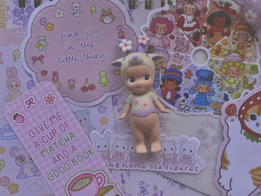 Photograph of my stickers and bookmarks with a purple/cream/pink Sakura sonny angel from my trinket collection. My products include a strawberry shortcake colour wheel sticker featuring eight fruit girls in each colour, a “find joy in the little things” flower sticker featuring trinkets, a Mofusand strawberry cat fan art sticker, “I bring home six figures” Sylvanian families fanart sticker, trinkets sticker sheet, and a “give me a cup of Matcha and a good book” bookmark