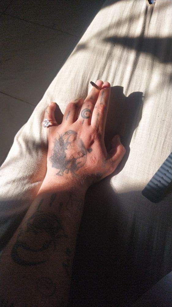 Foto de um braço tatuado pegando sol somente na mão que está segurando um cigarro e usando de acessório um anel de plástico e criança