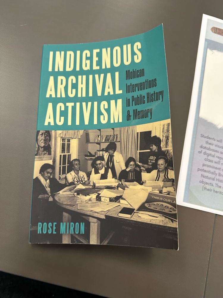 The cover of the book “Indigenous Archival Activism: Mohican Interventions in Public History and Memory” laying flat on a gray desk with the edge of a printed paper on the right side.