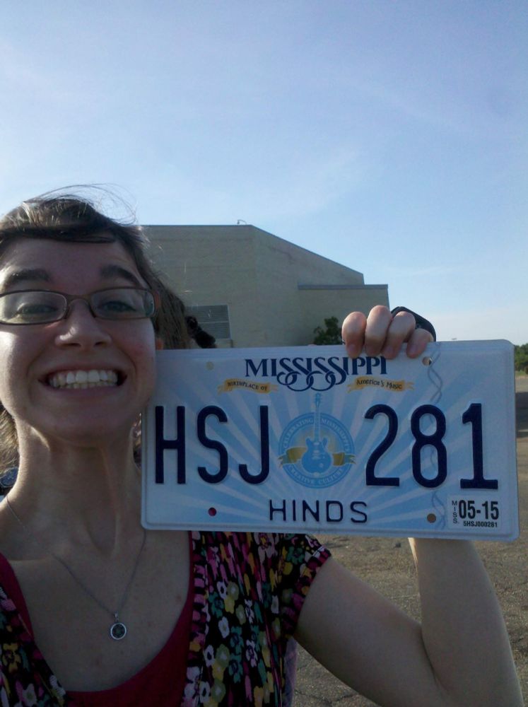 Woman with a big grin holding a license Mississippi license plate.