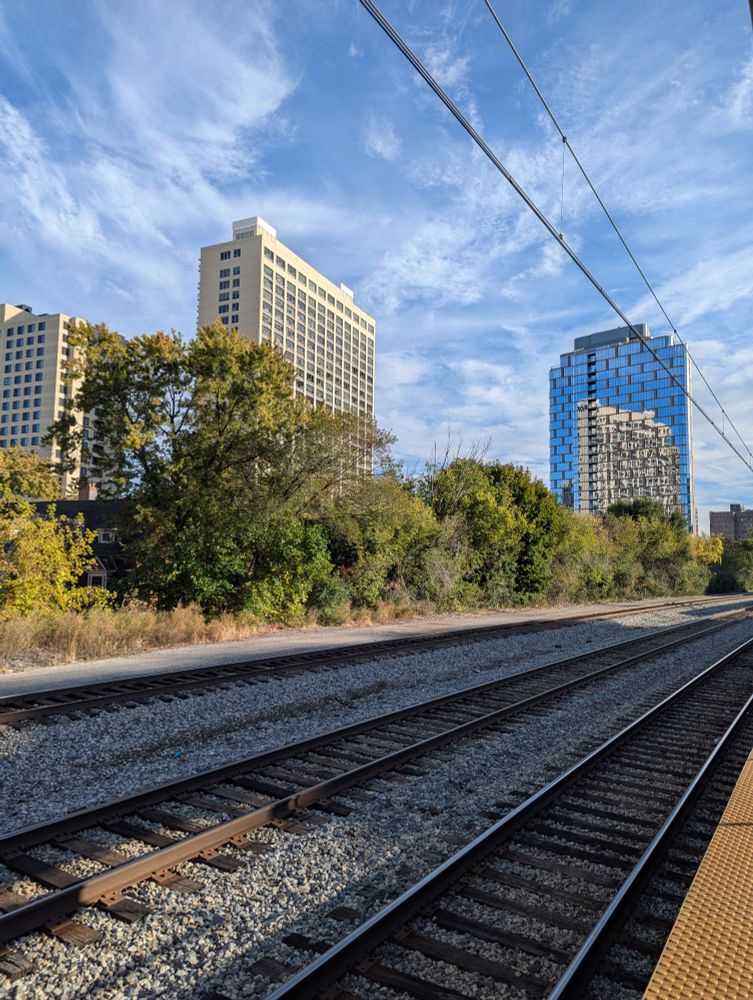 Behind three rows of train tracks is a tall, cream-colorer high-rise, the reflection of which is visible in the mirror-windowed highrise to the right. Power lines run through the top right corner of the photo. The sky is blue with plenty of wispy white clouds 