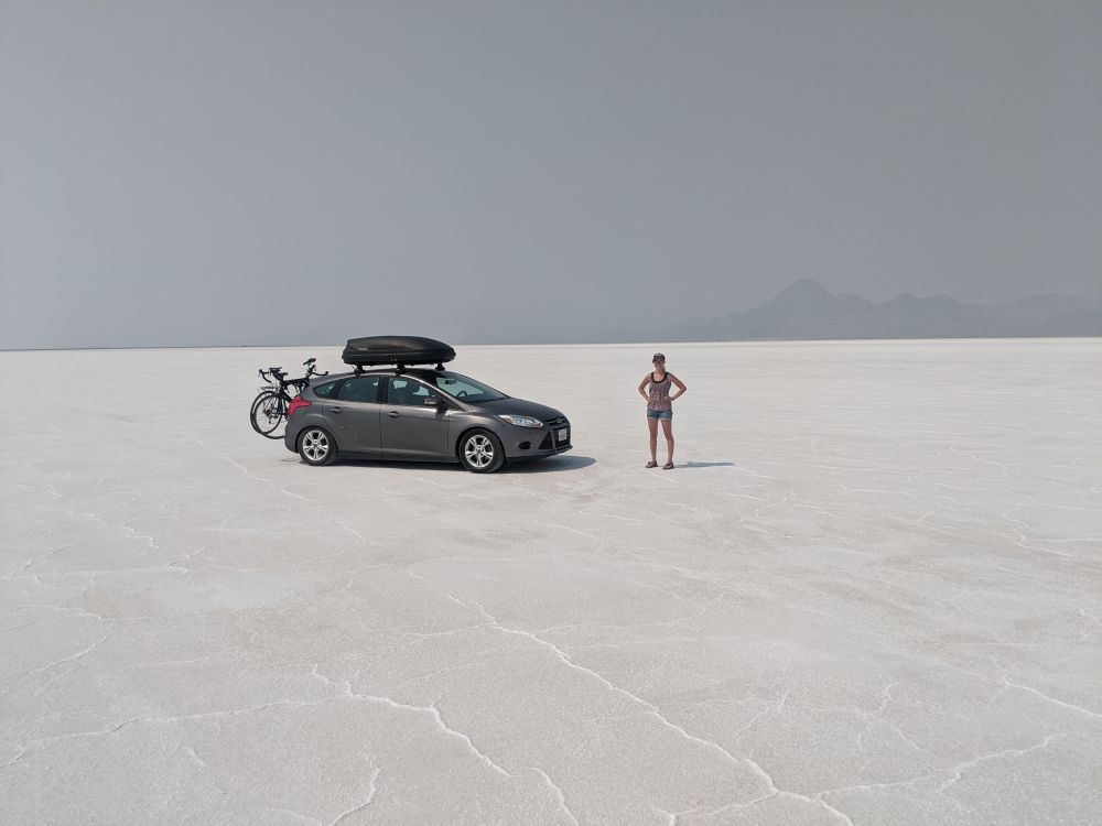 Gray car with bikes on back and cargo carrier on top. There is a woman in shorts next to the car. The car is on top of flat white ground (salt flats), and against a backdrop of a smoky/smoggy sky with mountains faintly visible in the background 