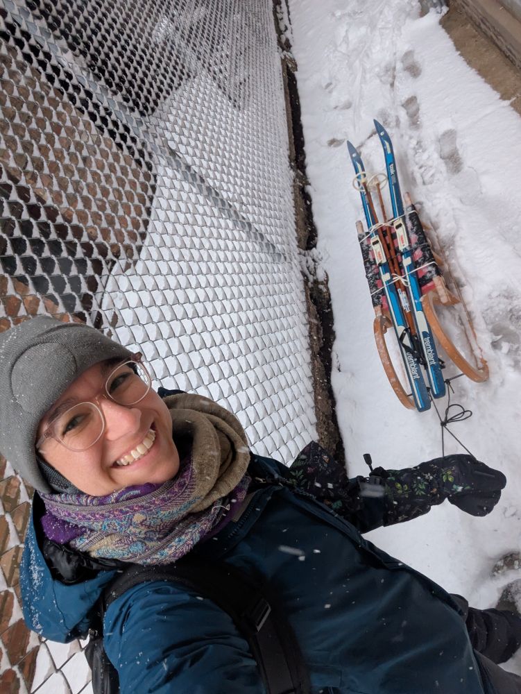 Woman wearing snow gear pulling an old-fashioned wooden sled, atop which are tied a pair of cross country skis and ski poles