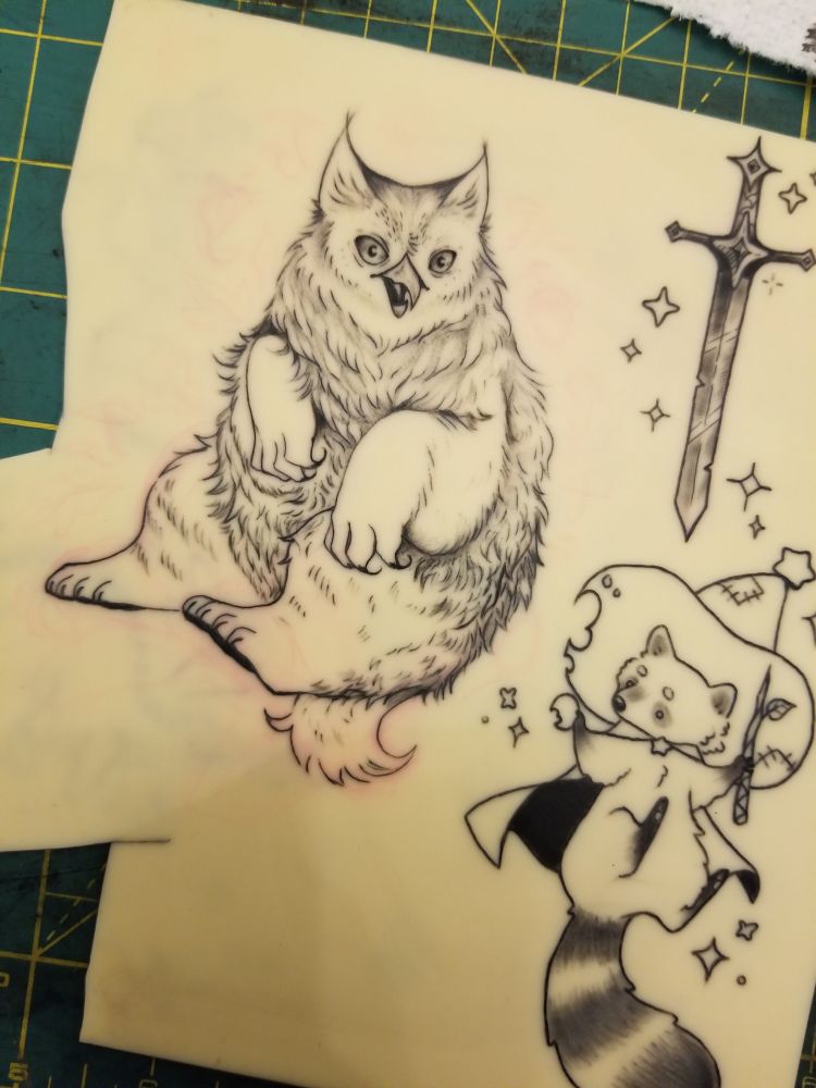 Photo of some fake skin with some practice tattoos on it! One is a fluffy owlbear, another is a doodle of a sword, and another is a little racoon mage. The owlbear and sword were freehand sketched on, the racoon was stenciled.