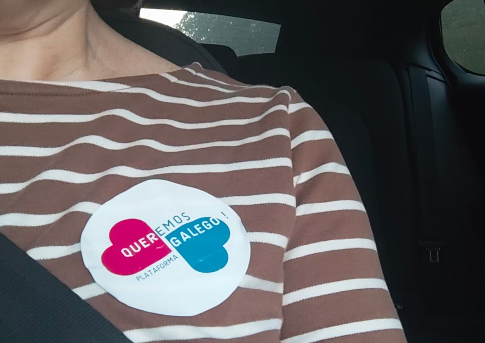 Un colante da Plataforma Queremos Galego! sobre a parte esquerda do peito dunha persoa que viaxa en automóbil. Só se ve o torso, cunha camisola marrón a raias brancas, o cinto de seguridade, e parte do habitáculo.