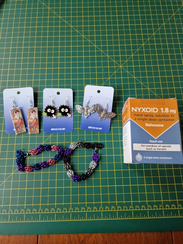 3 sets of dangle earrings (One of art by Alphonse Mucha, one of soot sprites from Ghibli's Spirited Away, and one of stylised metal death's-head hawkmoths), 2 chainmail bracelets in pride flag colours (bi and ace) and a box of 2 single doses of Naloxone.