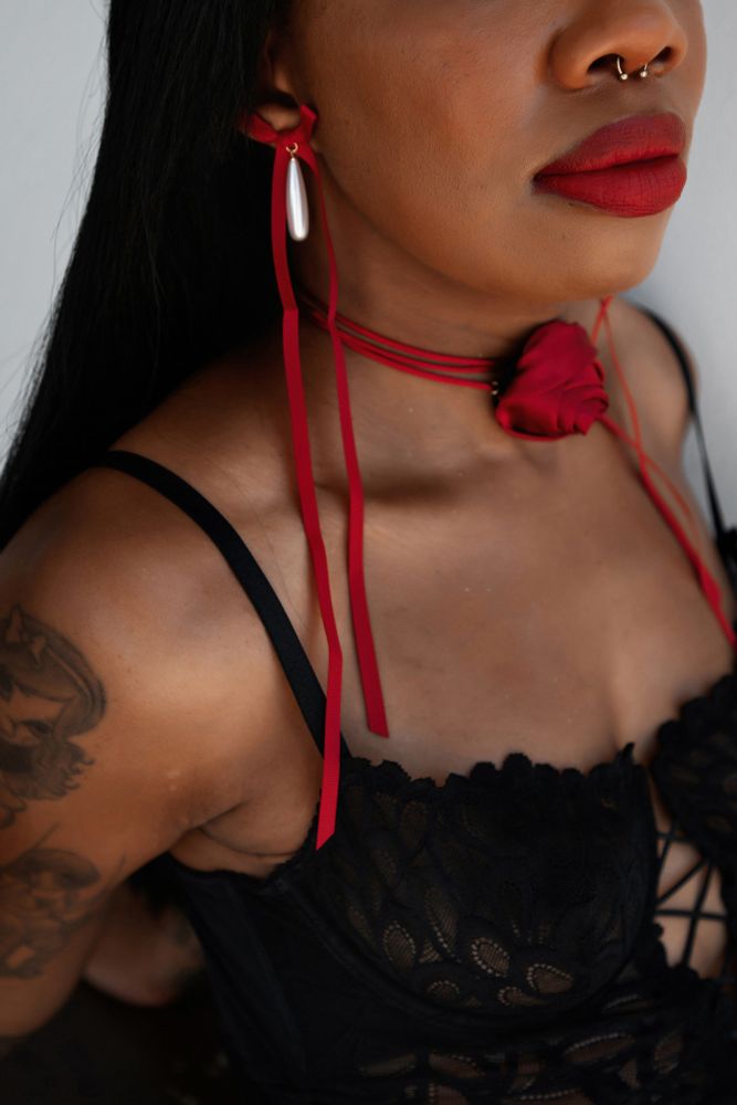 petit moments ad

a close up shot of dulces rose wearing a rosette bow pendant earrings and rosette tie around their neck. they have red lipsick and a septum. they are wearing a lace top. they hae long black hair and tattoos on their right arm.