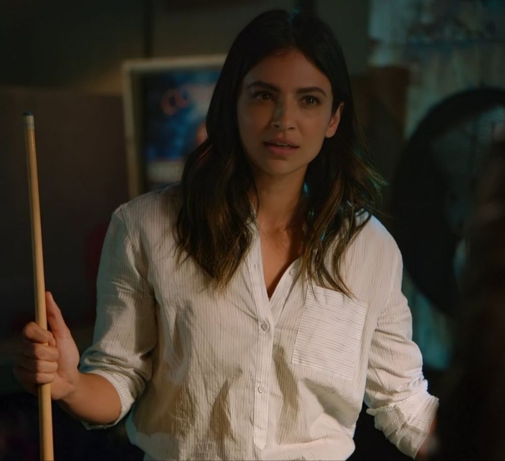 Floriana Lima as Maggie Sawyer in Supergirl. She is wearing a white button up shirt with the sleeves pushed up and holding a pool cue in her right hand. She has a questioning look on her face. 