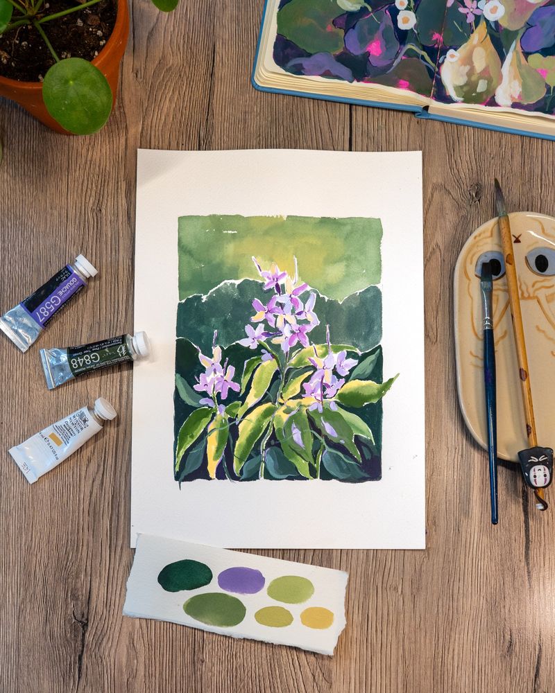 A watercolor painting of delicate lilac wildflowers with lush green leaves, placed on a wooden table. Surrounding the painting are paint tubes, brushes, a ceramic palette with a cat face, a sketchbook showing similar floral artwork, and a small color swatch card with greens and purples. A potted plant sits in the top left corner.