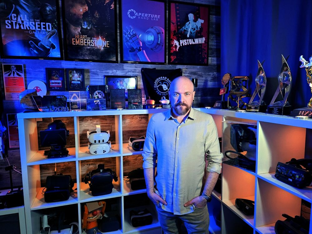 Image shows Denny Unger in his office standing in front of a display of VR headsets, awards, and posters featuring Cloudhead Games' IPs.