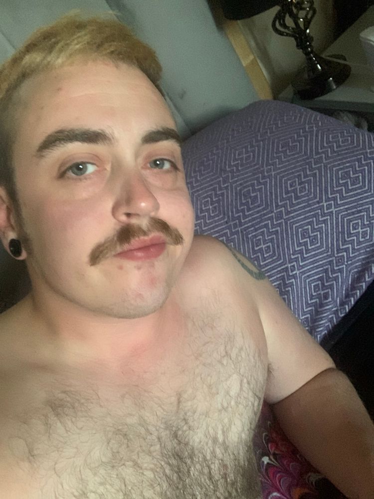 Jack is topless in bed. His hairy chest taking up most of the image. He gives a lopsided smile to the camera