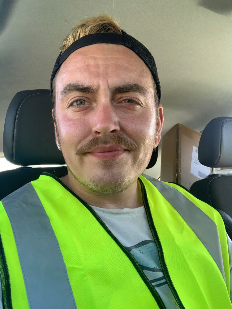 Jack sits in his car in a high vis jacket. Baseball cap in reverse. His skin is shiny with sweat. 