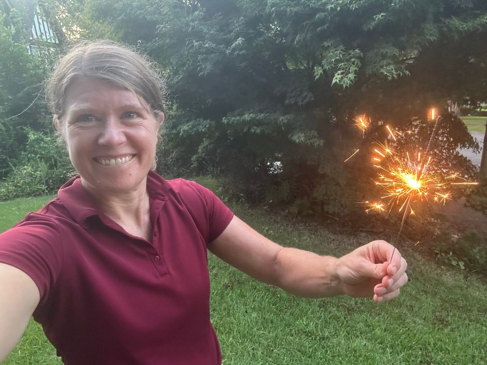 smiling blond woman in a red shirt stands in a lush green yard holding a lit sparkler in her left hand 