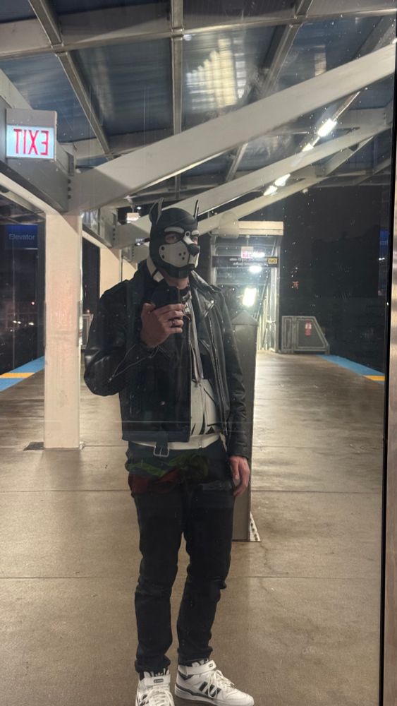 A person wearing a leather jacket and a pup hood on a train platform. 