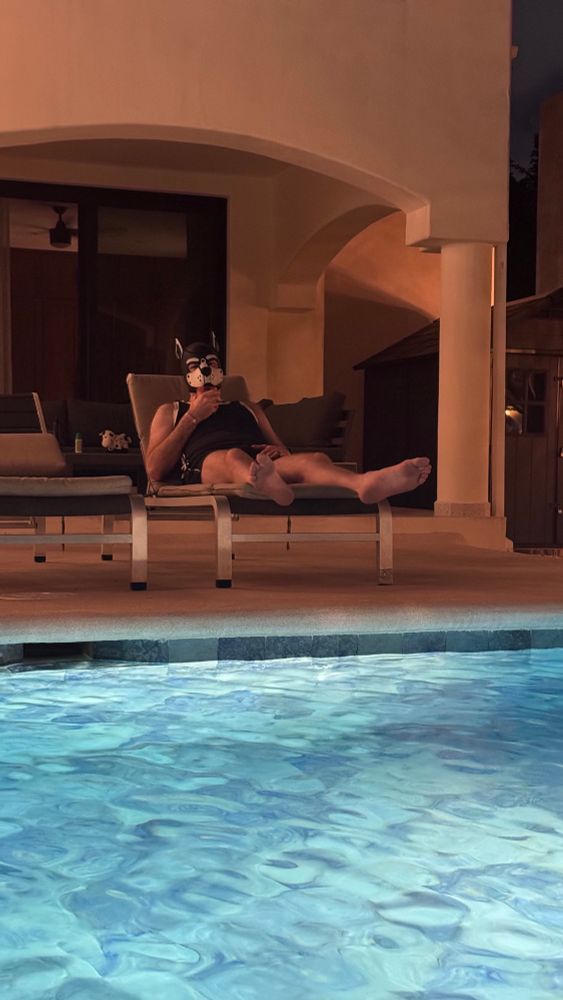 Looking across a pool, Tangent in a pool chair wearing his pup hood, a black tank top with white stripes on the side, and black shorts with a large resort house behind him. 