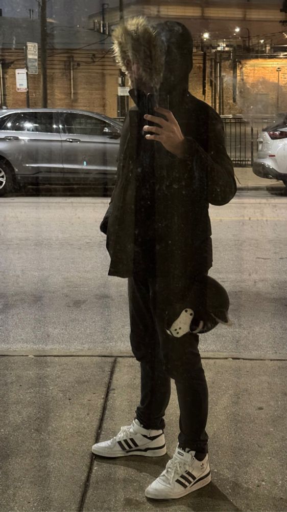 Person reflected in a mirror waiting for the bus wearing Adidas high tops and a pup hood on his belt. 