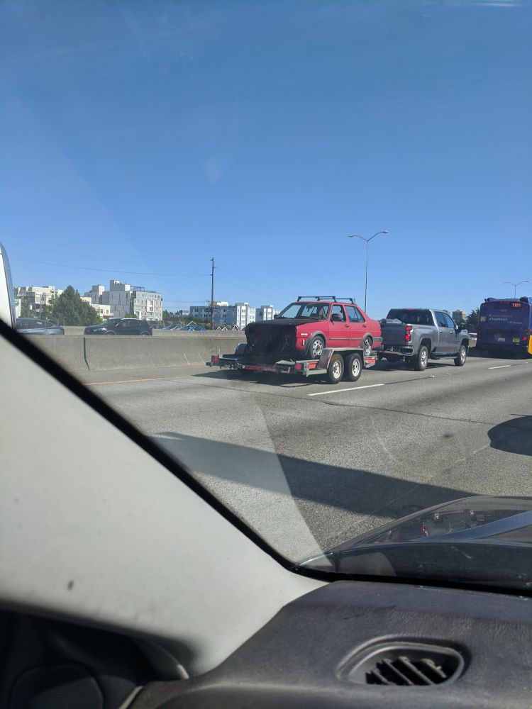A photo taken out a car window in traffic. A grey pick up truck hauls a trailer holding an older model red sedan with a black hood. The sedan is facing backwards, allowing the viewer to see that it has no front bumper or grill and in fact no engine. The bright sunlight casts a deep shadow making an only void of the empty space.