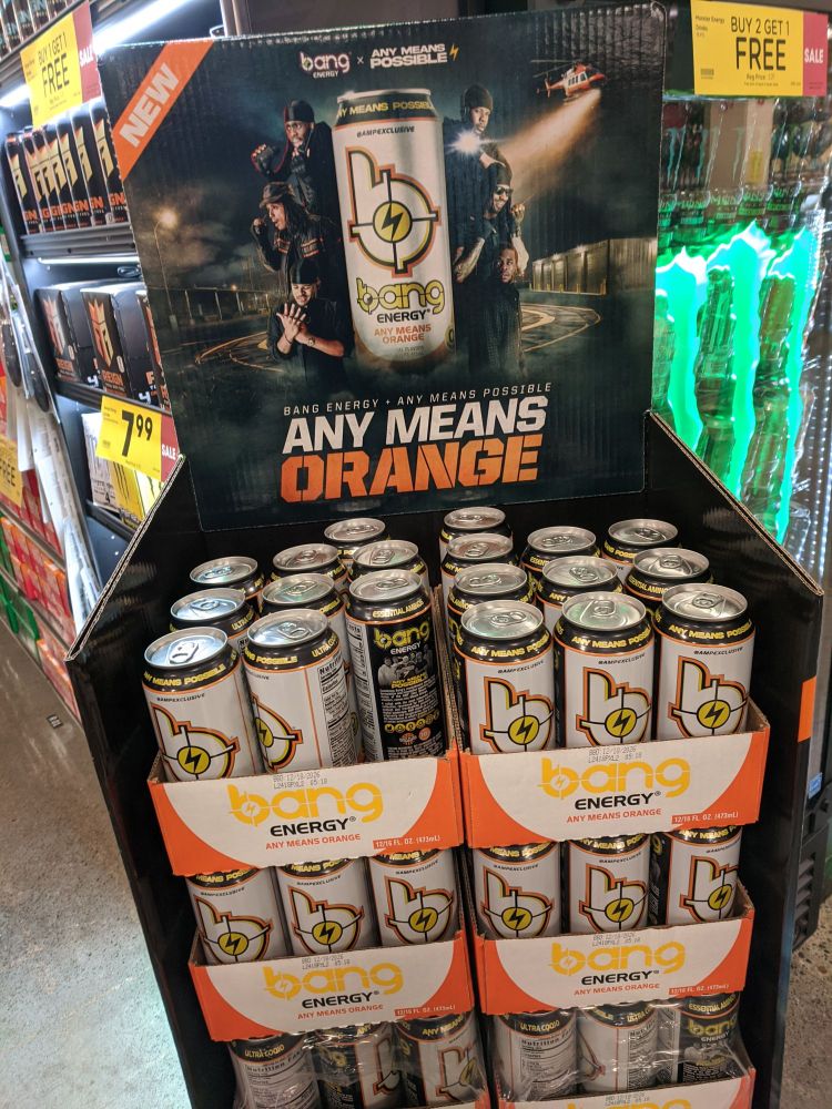 A cardboard grocery store display full of 'bang' brand energy drink cans. The cans are white with the logo of a lowercase b containing a stylized target with a bolt of electricity for a bull's eye. Above the cans the display stand has an advertisement for a flavor. A helpful banner at the top left tells us it's 'NEW'. The advertisement centers a can in a composition that may be intended as a reference/parody to heist movie posters. Six black men lean out from around the can, all dressed in black paramilitary-esque gear. Their poses and attitudes are all distinct, as though we are meant to infer personalities and perhaps even Heist Roles from this still. As a background the ad has a helicopter shining a search light into a landing zone at night. Along the top text says the brand name of 'bang' and then ANY MEANS POSSIBLE with the electrical bolt from the logo.
The name of the new flavor being advertised sits at the bottom of the image like a movie title. In big block white text it reads 'ANY MEANS', then on orange text 'ORANGE'.