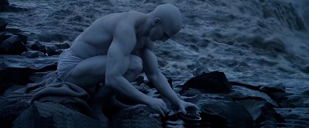 screenshot from prometheus (2012), a ripped bald man with corpse white skin and a loincloth on a stark icelandic beach 