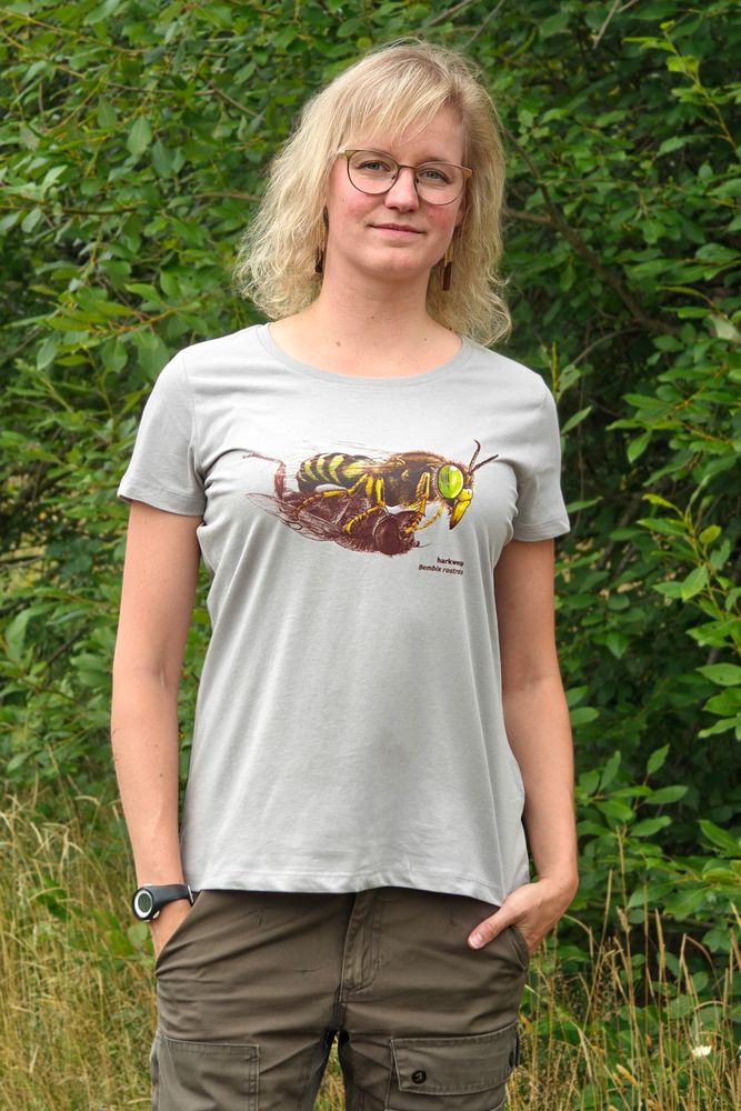 Een jonge vrouw met bril en halflang blond haar voor een onscherpe achtergrond van groene bomen. De vrouw kijkt naar de camera en draagt een grijs T-shirt, op de borst staat een kleurrijke harkwesp die een prooi draagt. De prooi is weergegeven als een lijnentekening in een warme bruine tint.