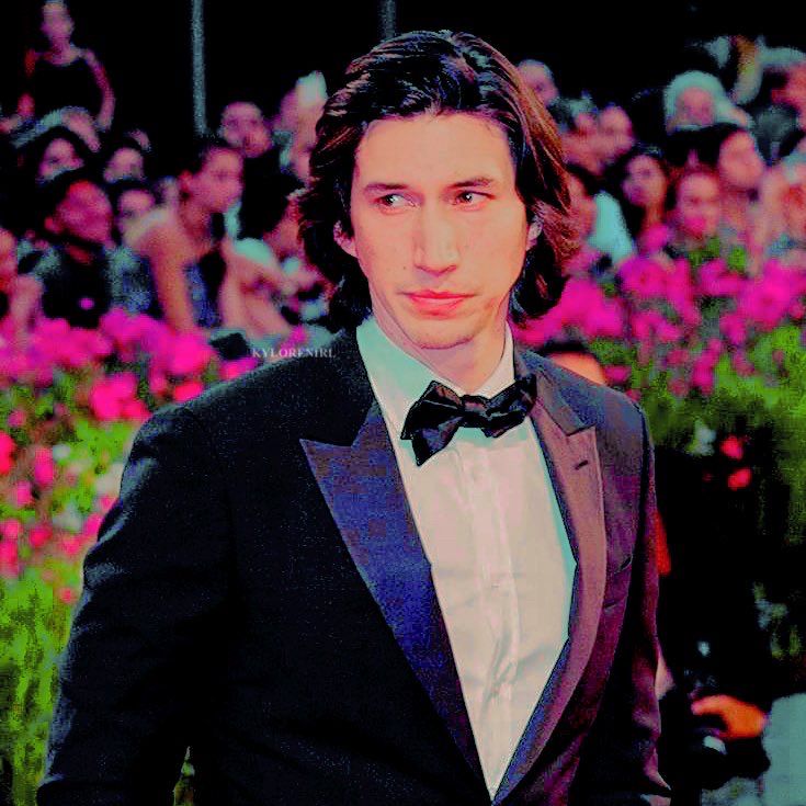 actor adam driver looking dapper in a tuxedo, behind him are rows of pink flowers and a crowd.