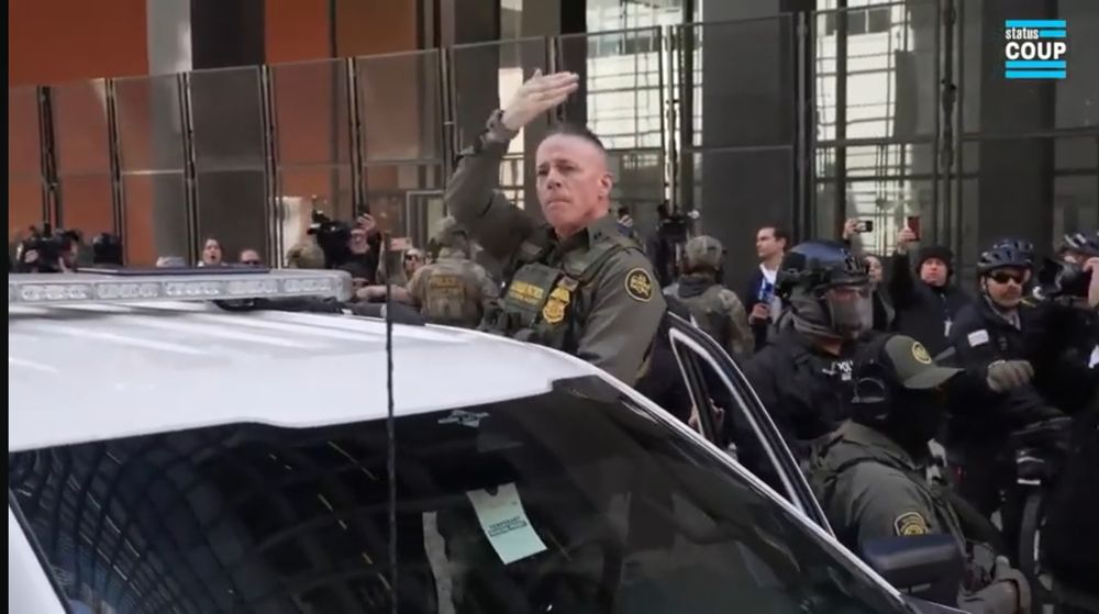 A short white male with sides of head shaved waves his right hand above his head. His fingers are tight together like a salute while his thumb is pointed up. He wears a dark green uniform with gold patches. He is standing between the open white car door and the driver seat. A crowd is behind him.