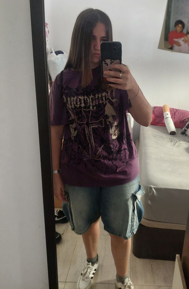 Yo en frente del espejo con una camiseta morada y unos pantalones cortos vaqueros. De fondo mi cama con un peluche con forma de cigarro.