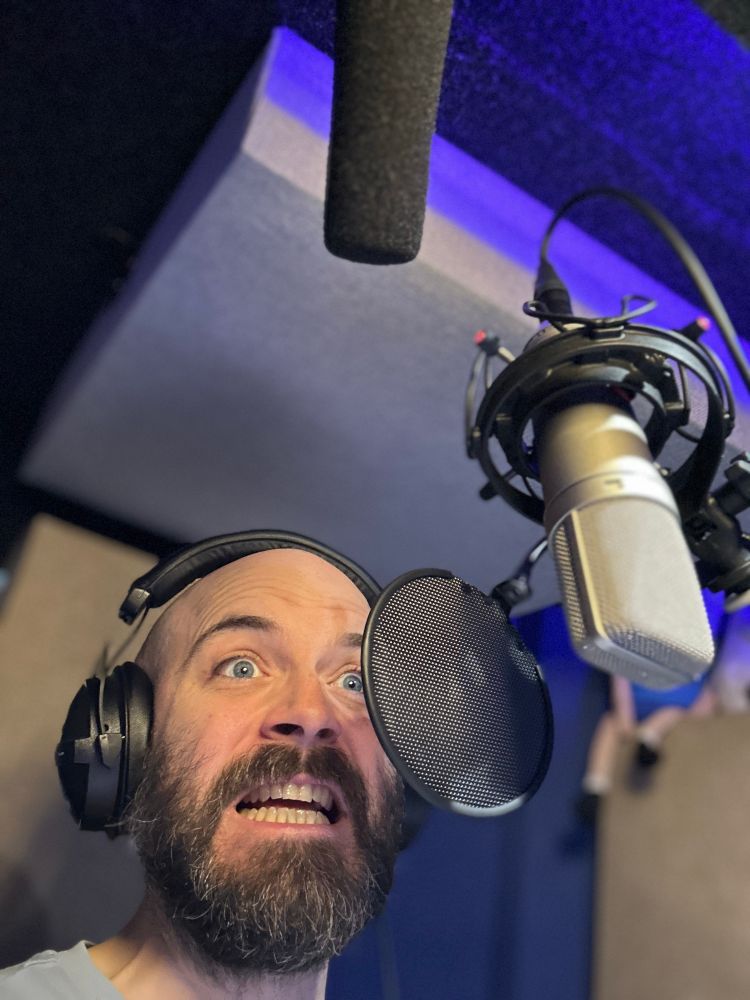 Photograph of a bald bearded man (me) overacting horribly in a sound booth. He wears headphones. There is a microphone in front of him. Please pit the microphone.
