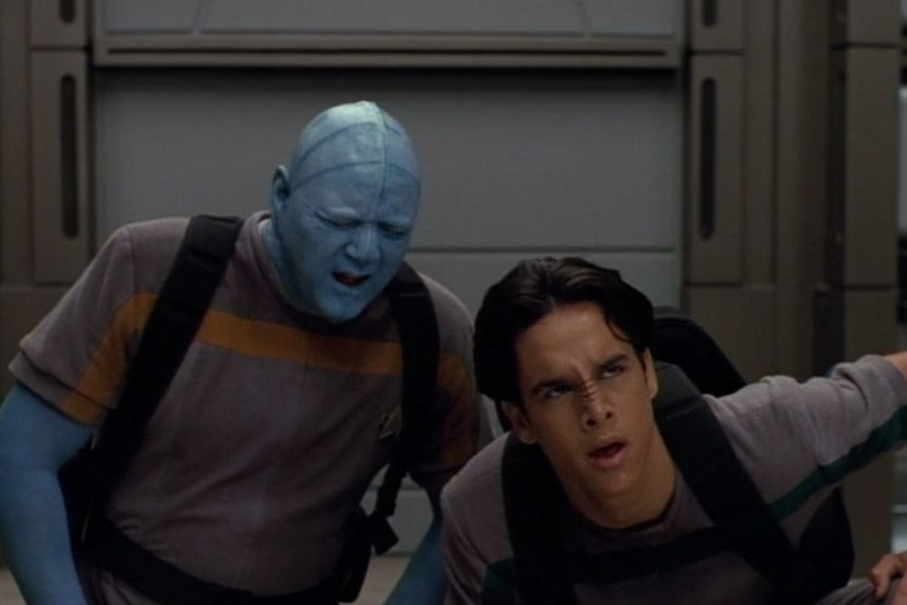 Derek McGrath as the Bolian Chell and Kenny Morrison as Bajoran crewman Gerron on the show Star Trek Voyager.  Bolian's are light blue skinned with horizontal darker blue stripes radiating from their ears across the top of their head with a generally humanoid shape with a seam along the midline of their entire body.  Bajorans are basically just humans with a ridged nose.  They're wearing matching t-shirts and backpacks looking tired.