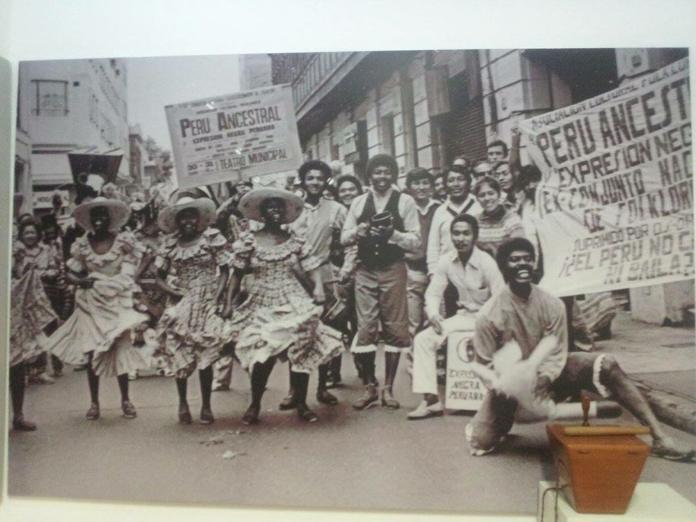 Black and white photo. Men and women of various indigenous, African or multihyphenate backgrounds dressed for a Peru Ancestral festival. 3 women of African descent at the front, in ruffled dresses and straw hats. Lots of smiling, happy faces.