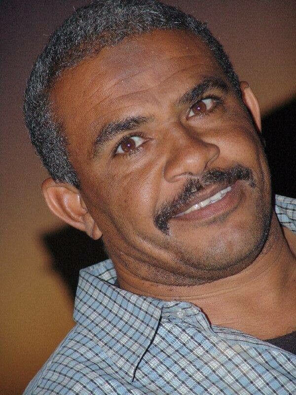 Tan to brown-skinned man with warn brown eyes, black hair beginning to grey ever-so-slightly. Thick and dark eyebrows and a small moustache framing his face.