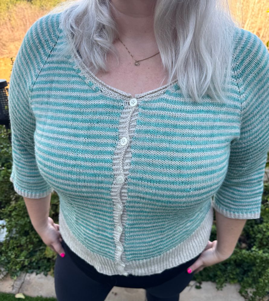 Hand knit cardigan on a woman standing outside. The cardigan is striped in beige and turquoise from yarn made of merino, alpaca, and silk. 