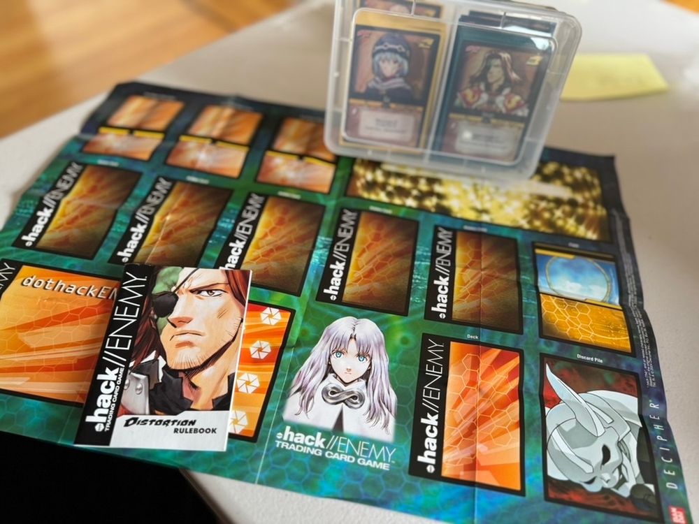 Two .hack//ENEMY Distortion precons in a plastic archival box, placed with the manual atop a paper playmat.