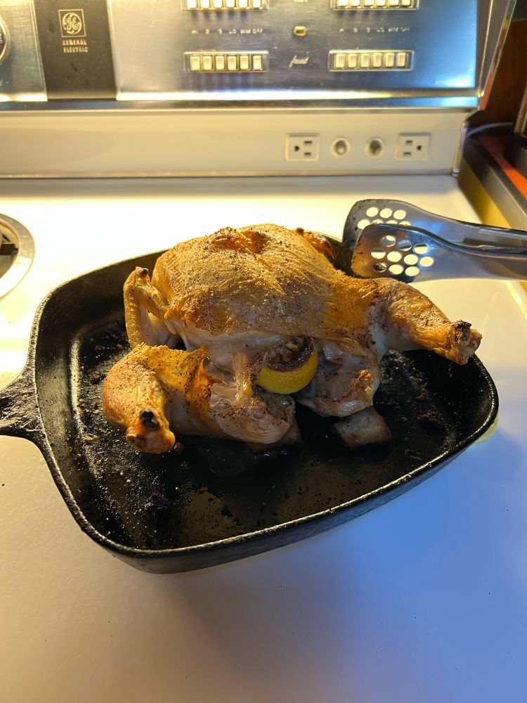 a roasted chicken in black cast iron skillet. there is a lemon stuffed inside the chicken. metal tongs are visible in the upper right quadrant of the photo. 