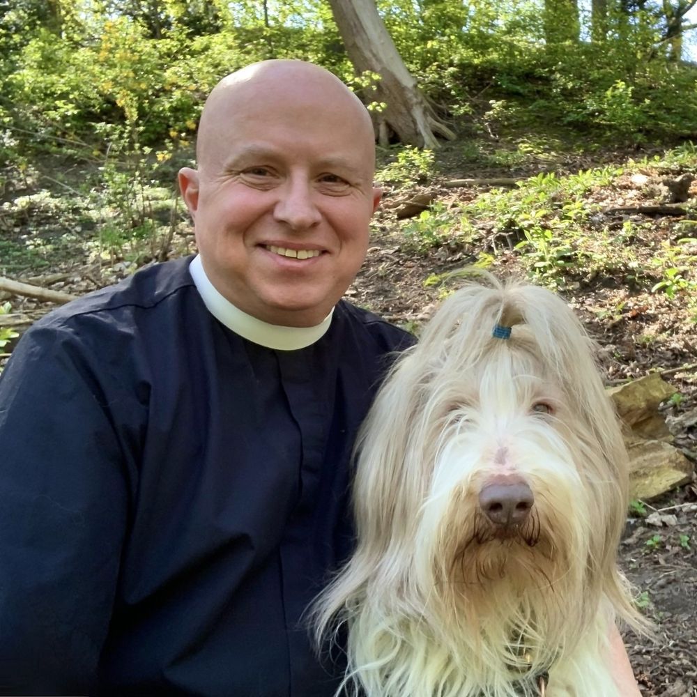 friendly priest in clerical collar kneels beside whitish bearded collie. both are facing the camera. background shows a lovely wooded trail.